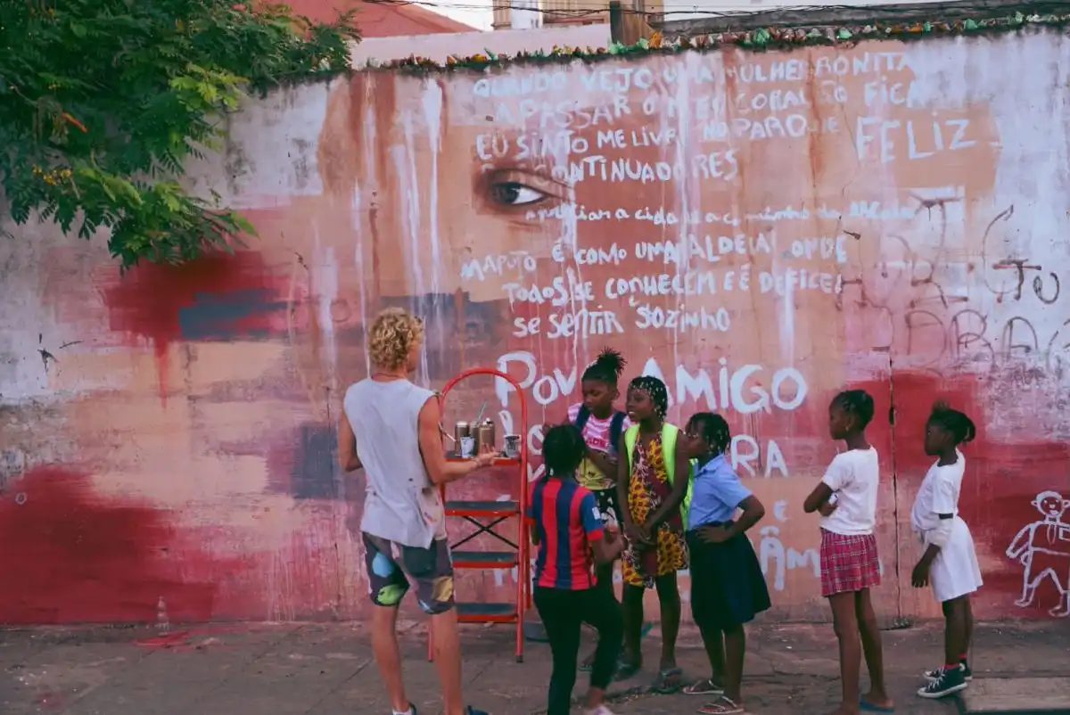 A person painting a mural on a wall, surrounded by children observing. The mural features an eye and Portuguese text.