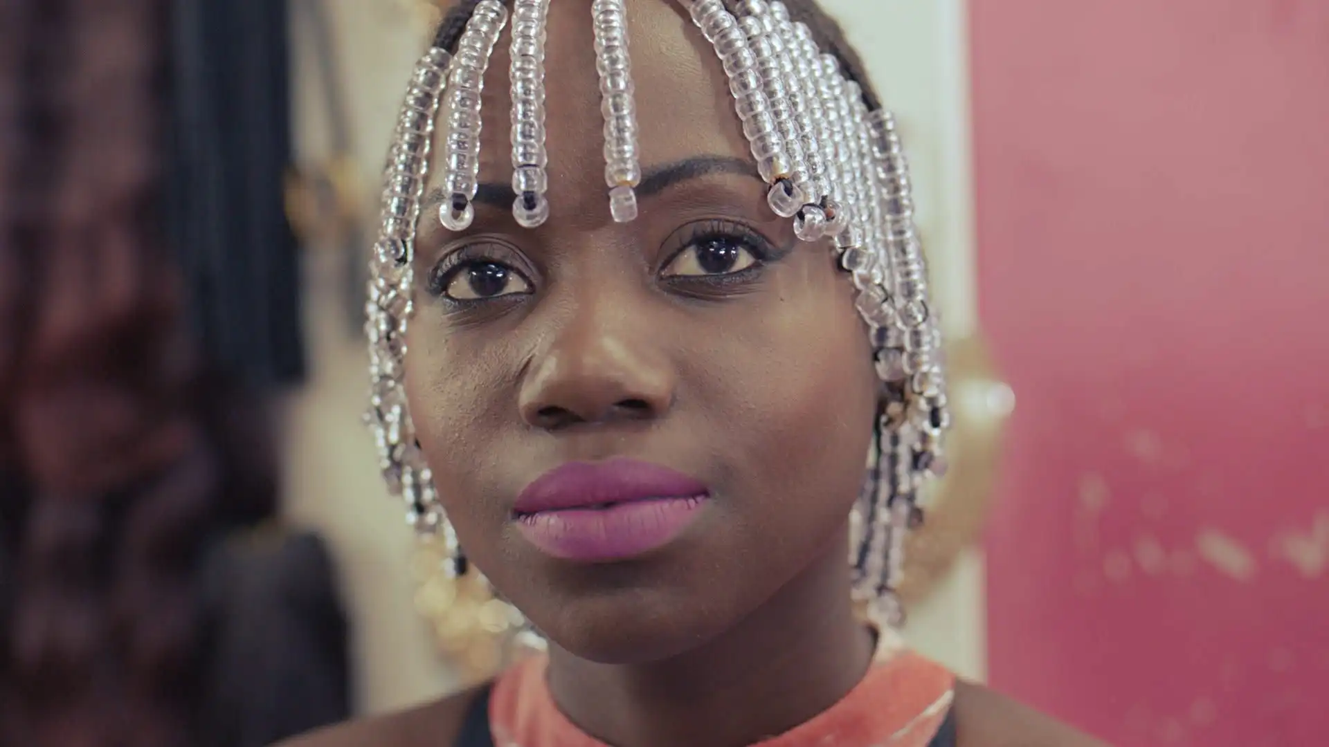 Woman with beaded hair and pink lipstick, wearing a coral top, looking thoughtfully to the side.