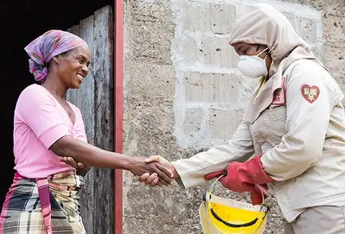 Two women shake hands outdoors; one wears a headscarf, the other protective gear with a mask, gloves, and a hard hat.