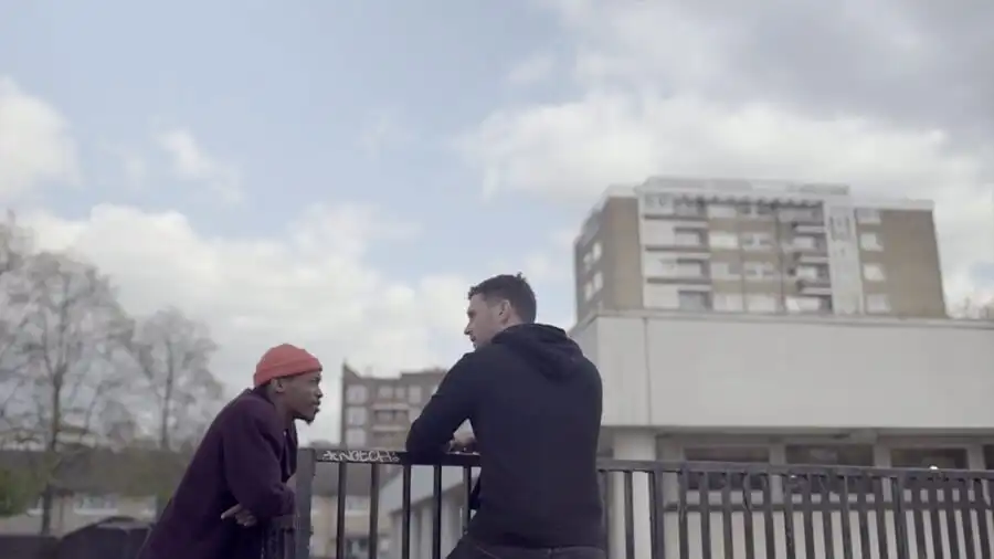 Two people chatting casually by a railing, with a cityscape and cloudy sky in the background.