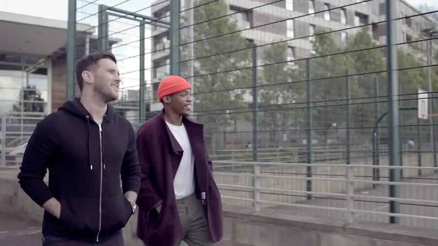 Two people walking and talking near a fenced sports area, one wearing a black hoodie, the other a red beanie and maroon coat.