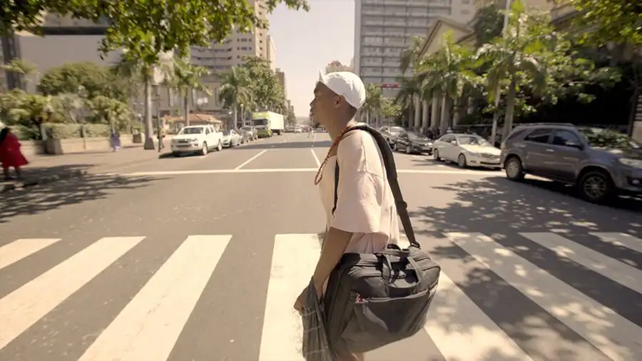 Person crossing a city street at a crosswalk, carrying a large bag, with trees and buildings in the background.