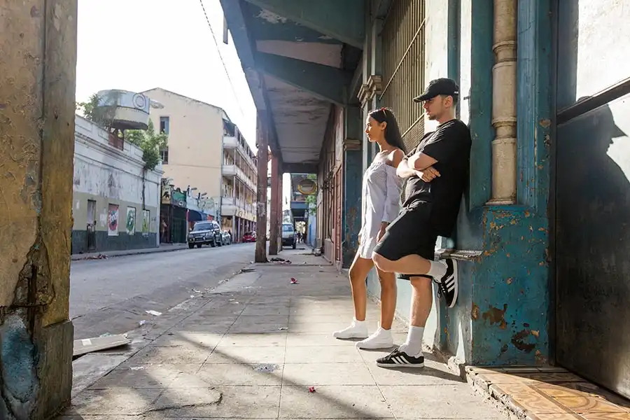 Two people stand against a blue building on a quiet, sunlit street lined with old structures and scattered debris.