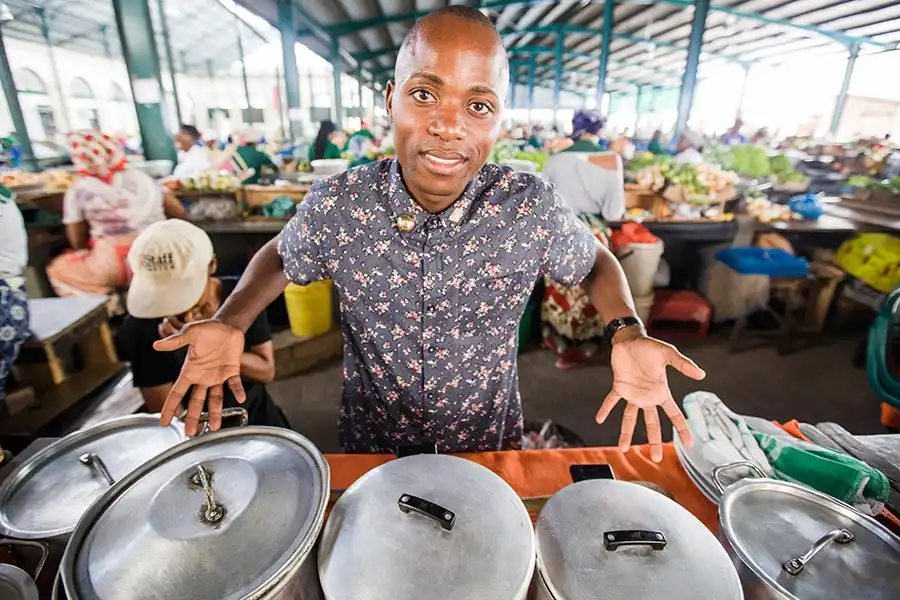 Person standing behind a market stall with covered pots, smiling, in a busy indoor market setting.