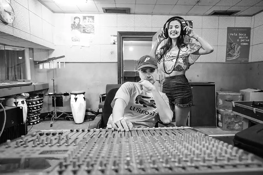 Two people in a recording studio. One sits at a mixing console, while the other stands behind wearing headphones, smiling. Black and white photo.