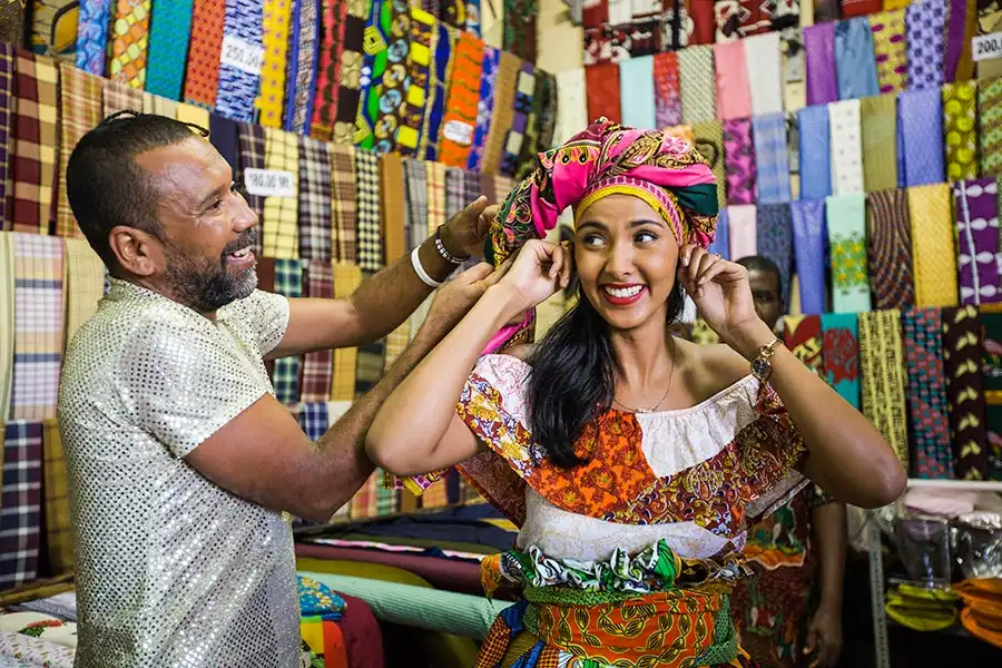 A woman smiles as a man helps her try on a colorful headwrap in a vibrant fabric store.