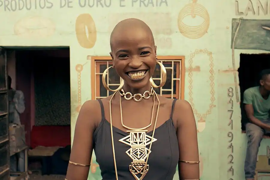 Smiling person with bold jewelry and shaved head, standing in front of a colorful building.