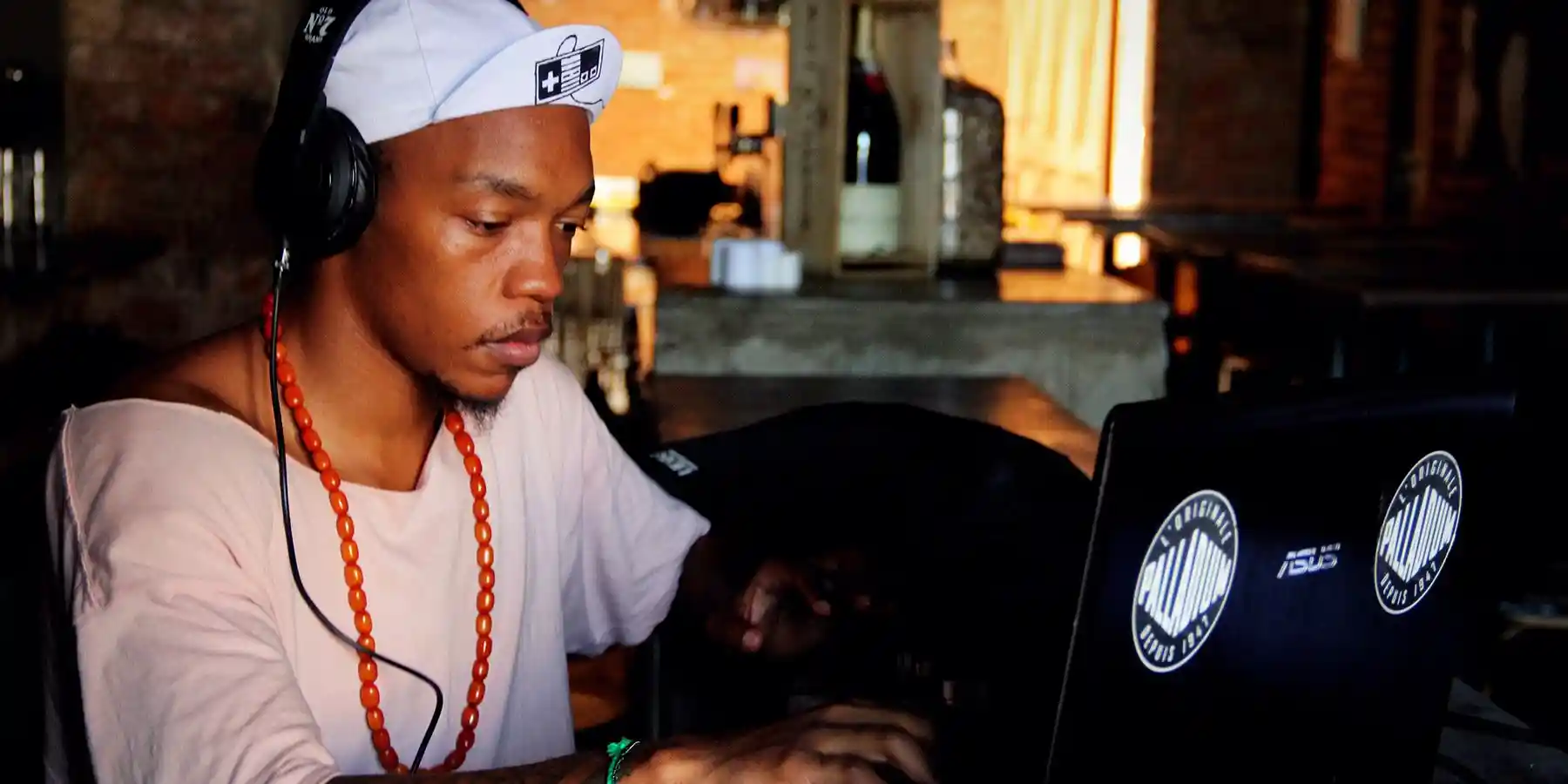 Man in a white cap and headphones working on a laptop in a cozy, dimly lit room.