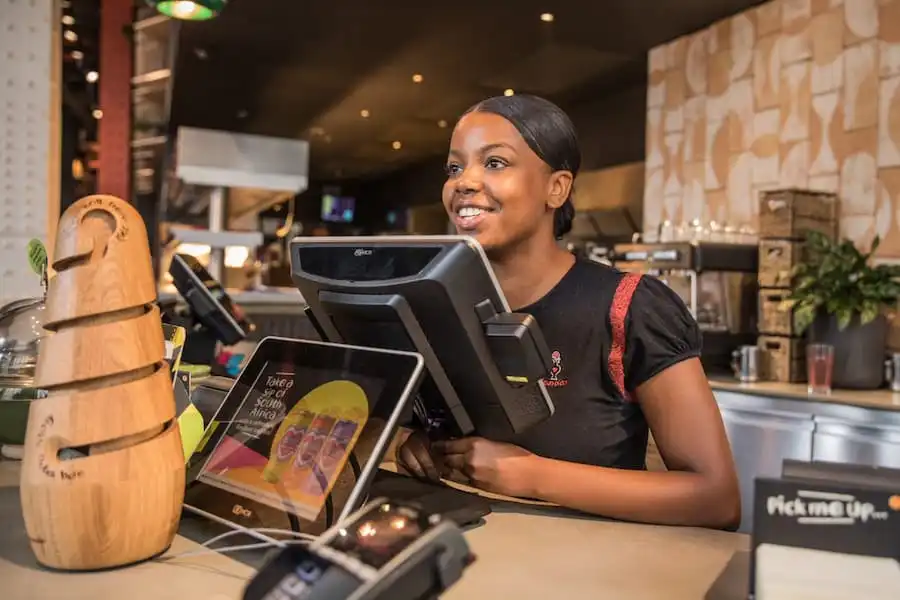 Smiling employee at a Nando's counter, ready to assist customers. Menu items and promotional display visible in the background.