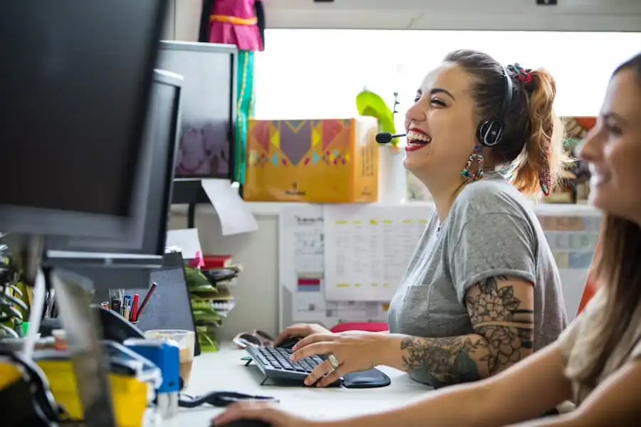 Two women at a desk, smiling and wearing headsets, working on computers in a bright office space.